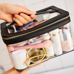 Truffle clear cosmetic bag trimmed in black leather, held by a hand over a tiled bathroom wall. The bag is filled with skincare and makeup items.