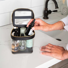Truffle Clarity Mini Train Case, trimmed in black leather, filled with beauty items sitting on a bathroom counter.