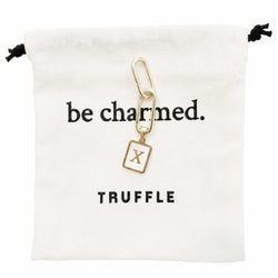 Truffle gold letter charm laying on top of a cotton pouch that says be charmed. 