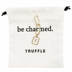 Truffle gold letter charm laying on top of a cotton pouch that says be charmed. 
